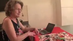 Reife Frau mit Brille und Locken steht lächelnd in der Küche neben einem Laptop auf rotem Tisch
