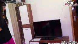 Schwarzhaarige Schönheit im schwarzen Top steht neben einem großen TV-Möbel im Wohnzimmer und lächelt verführerisch.
