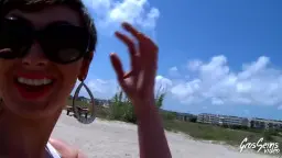 Frau mit Sonnenbrille lacht am Strand, Hand winkt in die Kamera