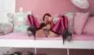 Lara liegt in pinkem Zimmer auf weißem Sofa, trägt nur schwarze Strapse und High Heels, spreizt die Beine und hält einen Dildo in der Hand.